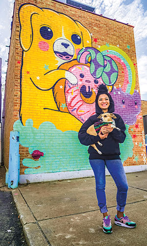A person holding a dog in front of a brick wall painted with a huge cartoon dog holding a donut